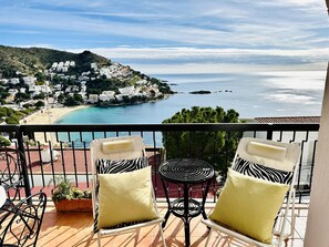 Een balkon met twee stoelen en een tafel, met uitzicht op een kustplaatsje met gebouwen en een strand.