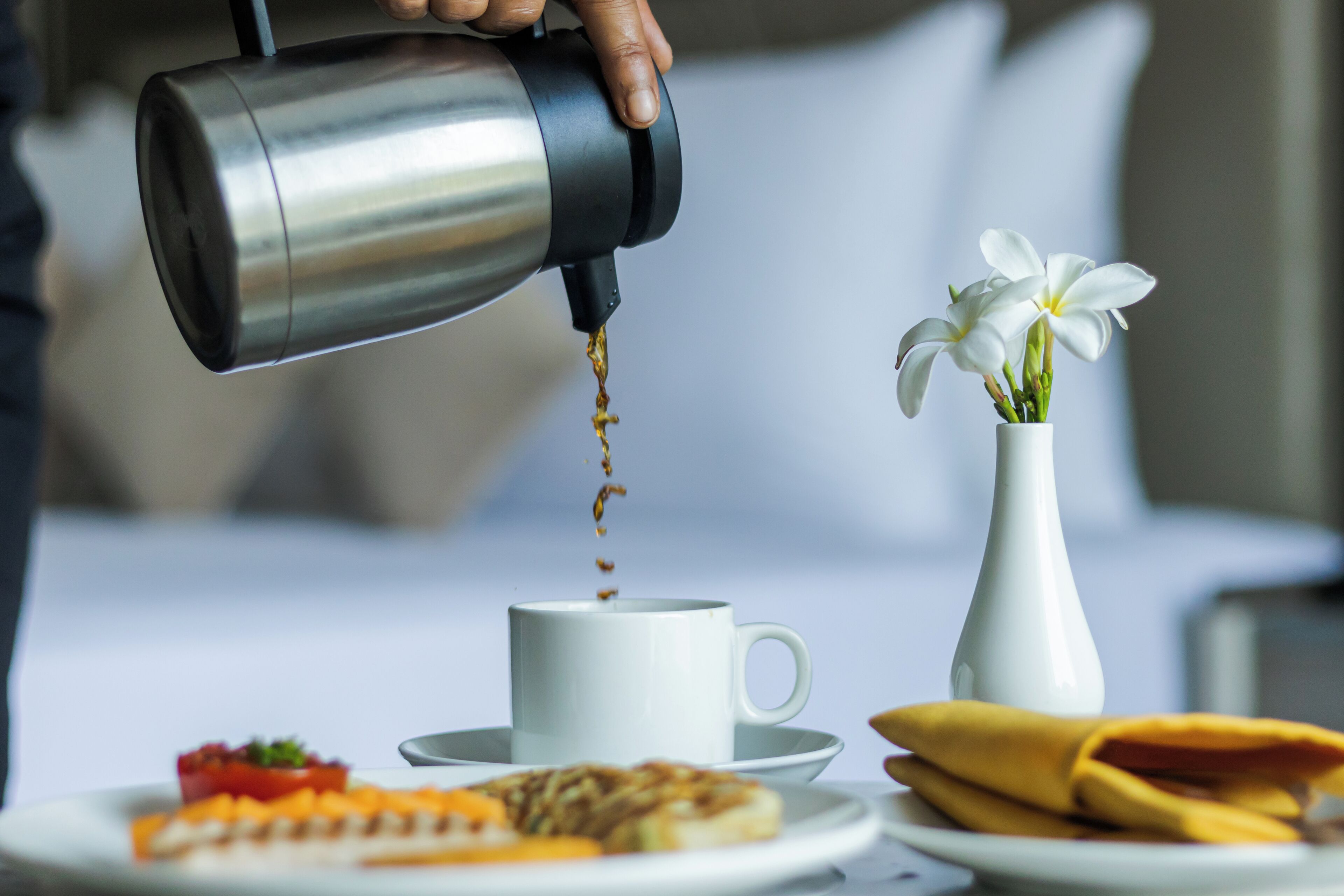 Una mano vertiendo café en una taza, un jarrón blanco con flores y un plato de comida.