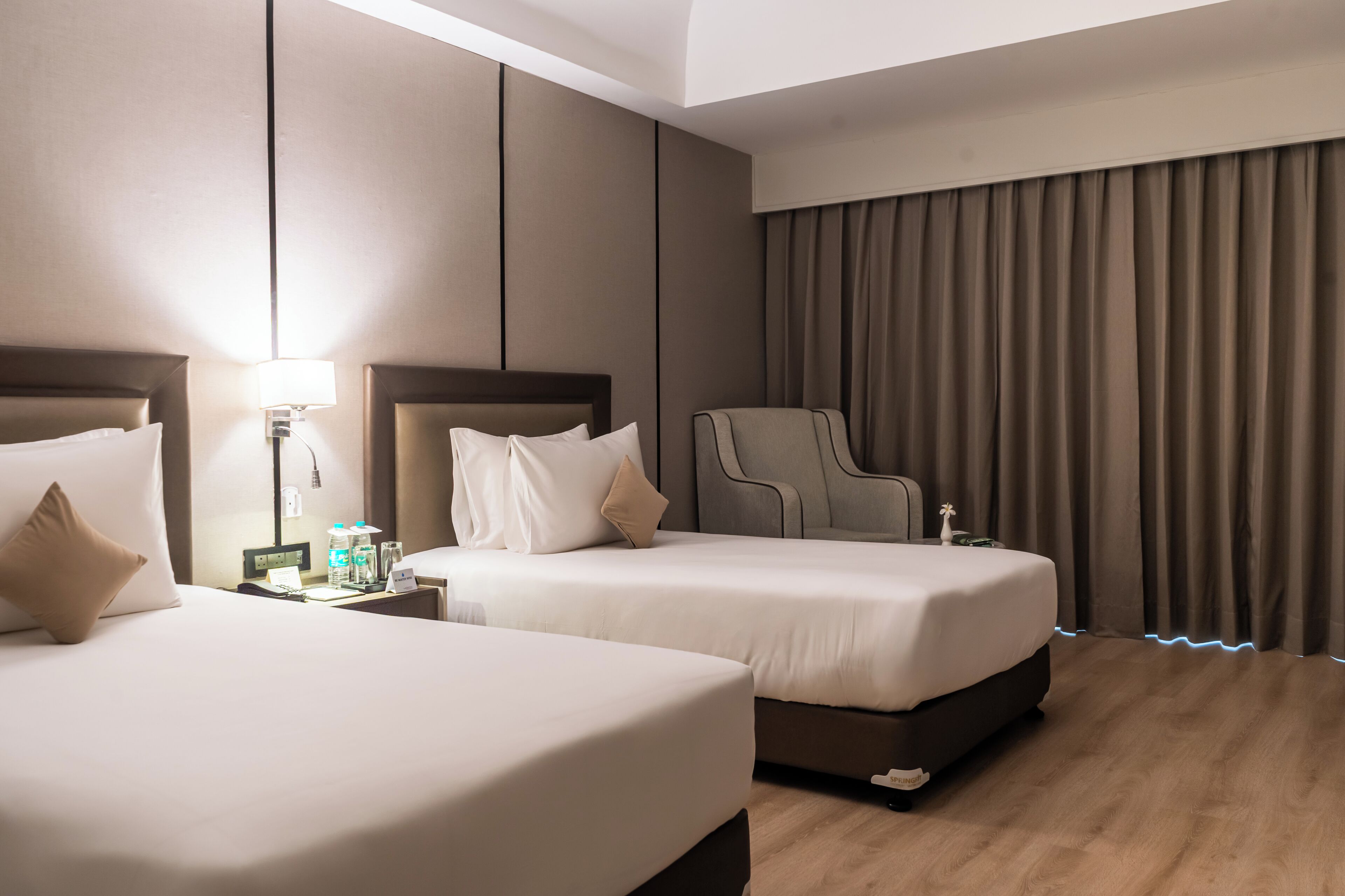 A hotel room with two beds, a nightstand with a lamp, a chair, and a large window with curtains.