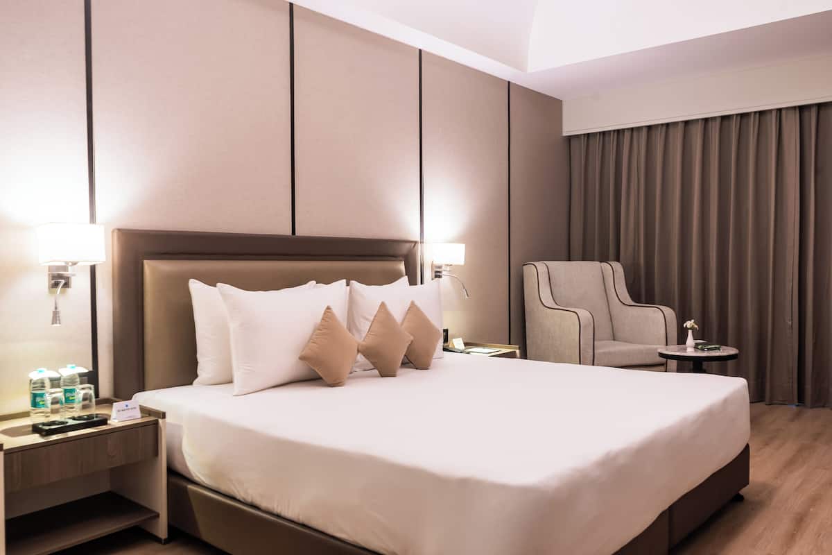 Ein modernes Hotelzimmer mit einem großen Bett, einem Sessel, einem kleinen Tisch und einem Nachttisch mit Lampe und Wasserflaschen.