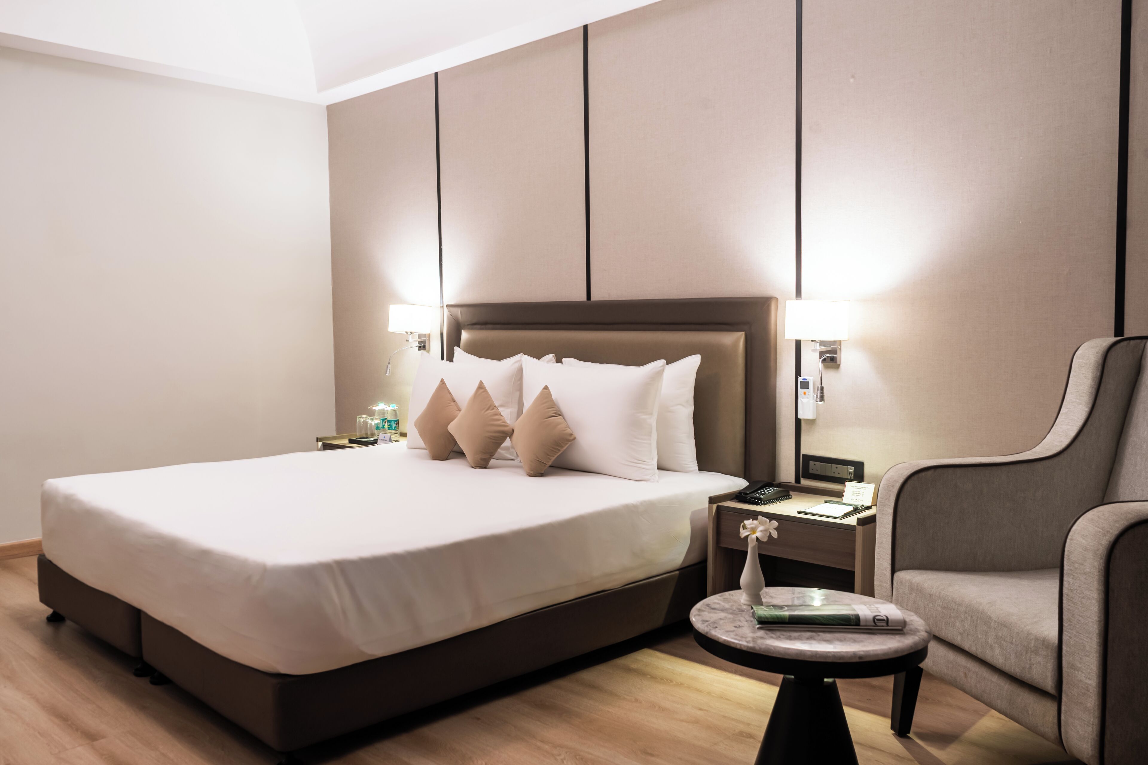 A hotel room with a large bed, a nightstand with a lamp, a chair, and a small table with a book.