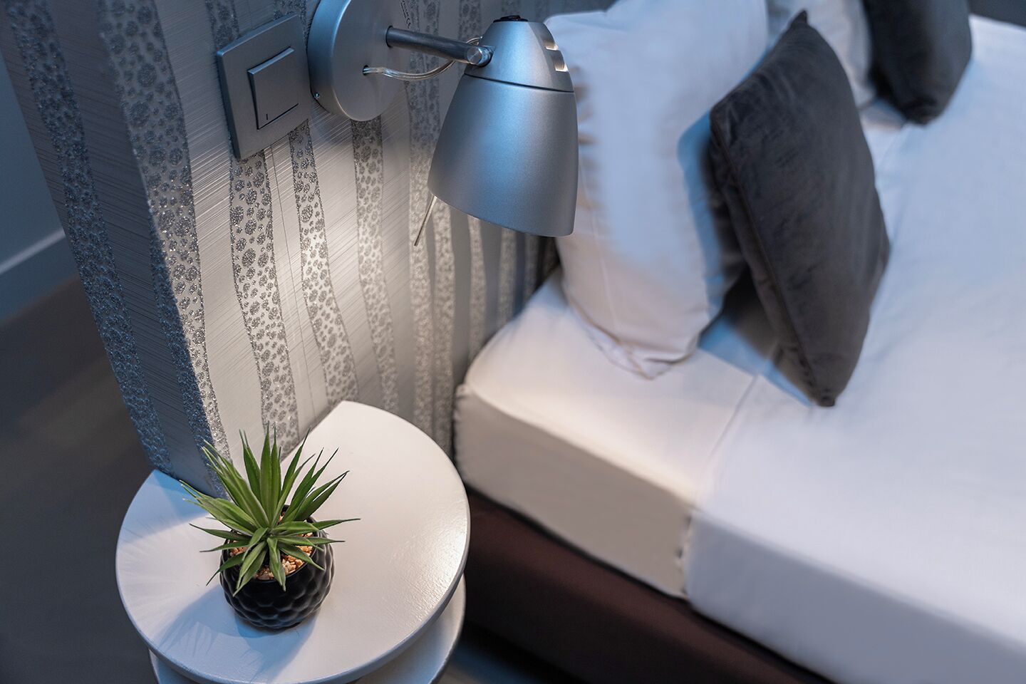 A neatly made bed with pillows, a bedside lamp, and a small potted plant on a round table beside the bed.