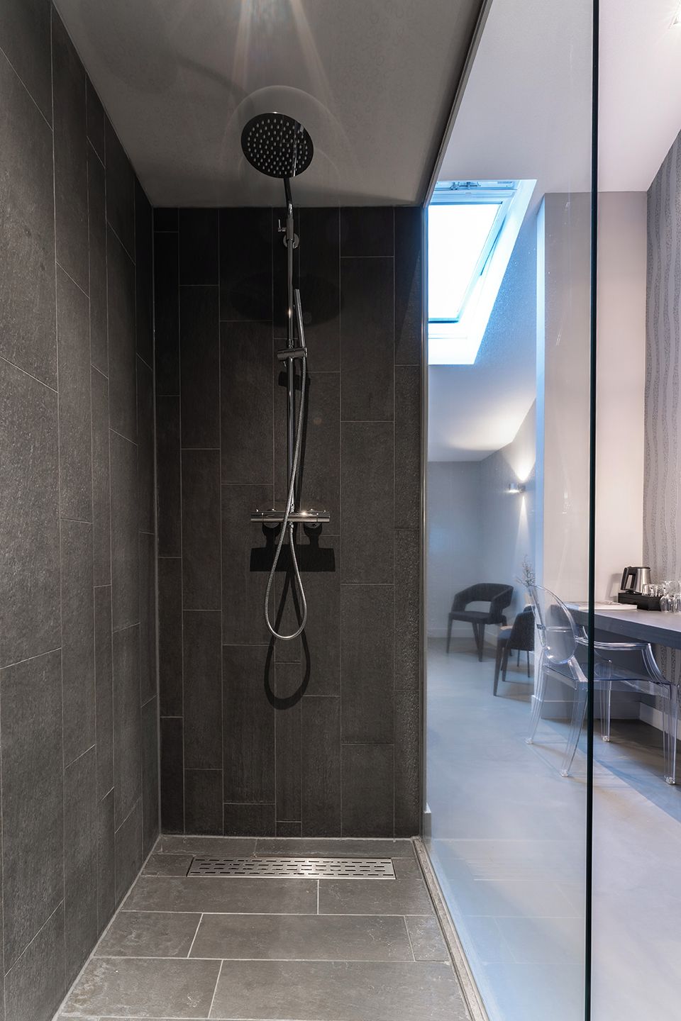 Eine moderne Dusche mit Oberlicht, dunklen Fliesen und einer Glaskabinentür, die in einen Essbereich führt.