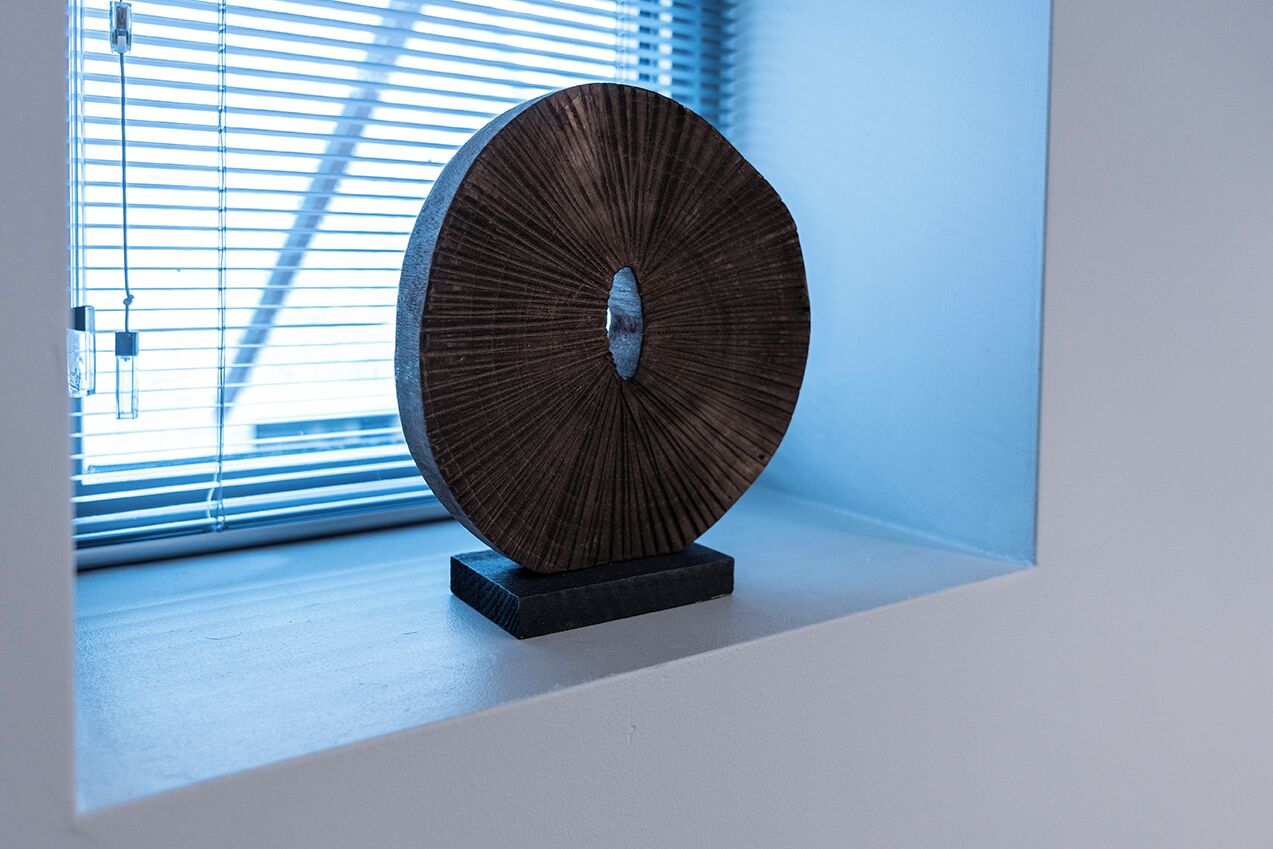 Una escultura circular de madera sobre una base oscura, colocada en un alféizar con persianas en el fondo.