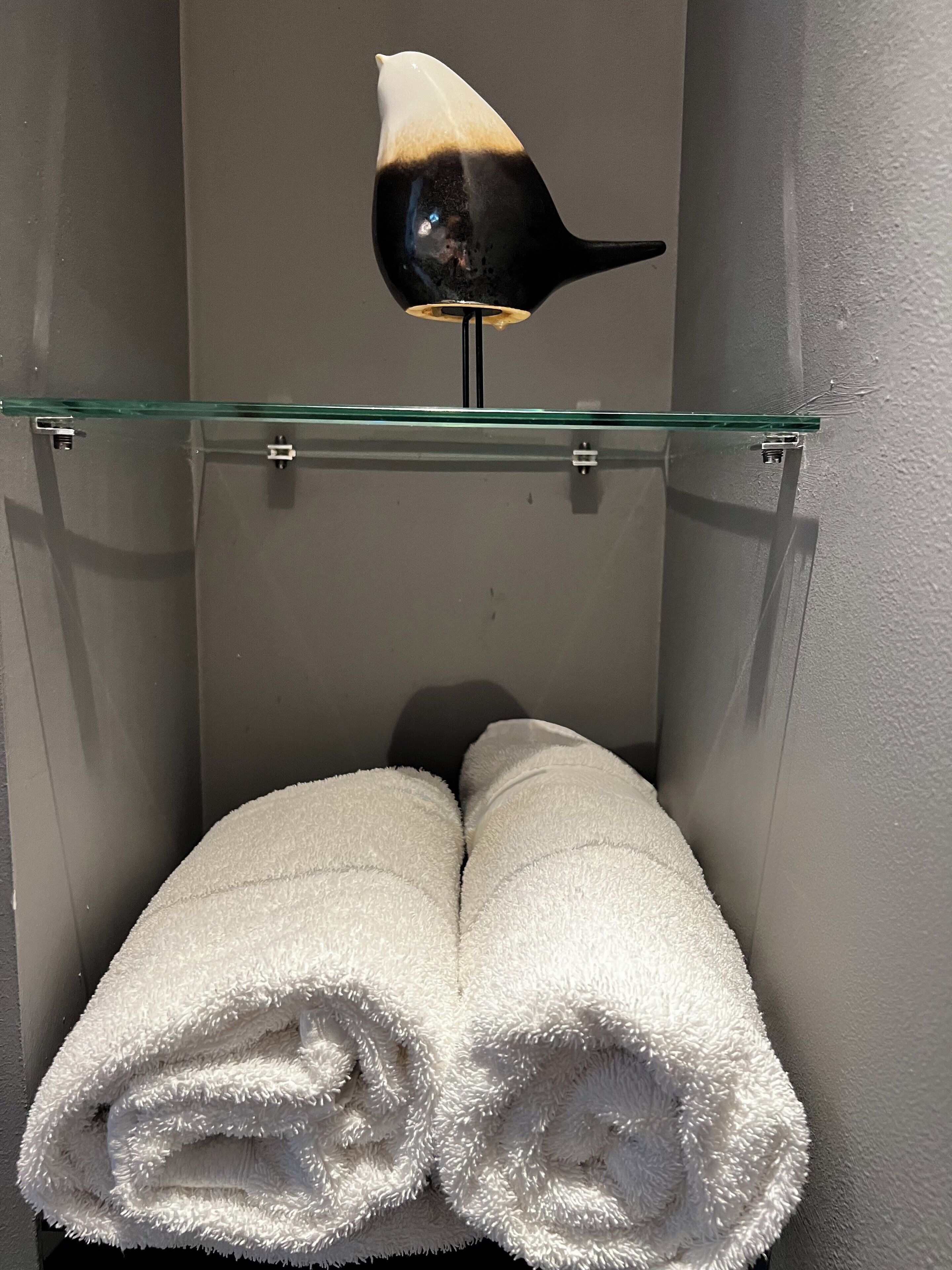 A bathroom with a glass shelf holding a black and white object, a white towel rolled up, and a white fluffy towel.