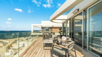 Una terraza moderna al aire libre con vista al mar, amueblada con sillas y mesas de madera y una barandilla de vidrio.