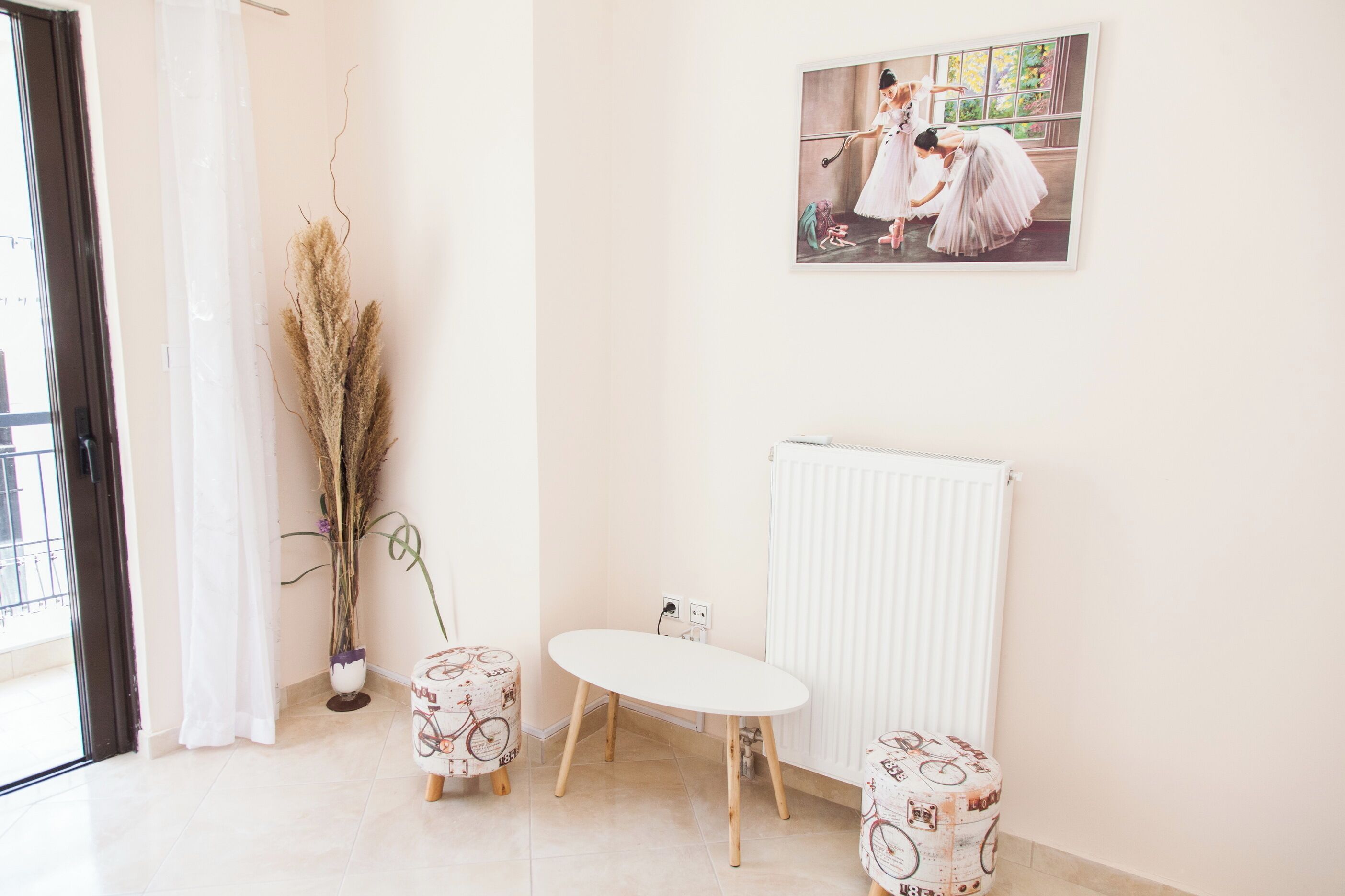 A room with a white radiator, a white table, two patterned stools, a potted plant, and a framed picture on the wall.