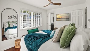 A bedroom with a bed, a ceiling fan, a window with blinds, a mirror, and a wall-mounted TV.