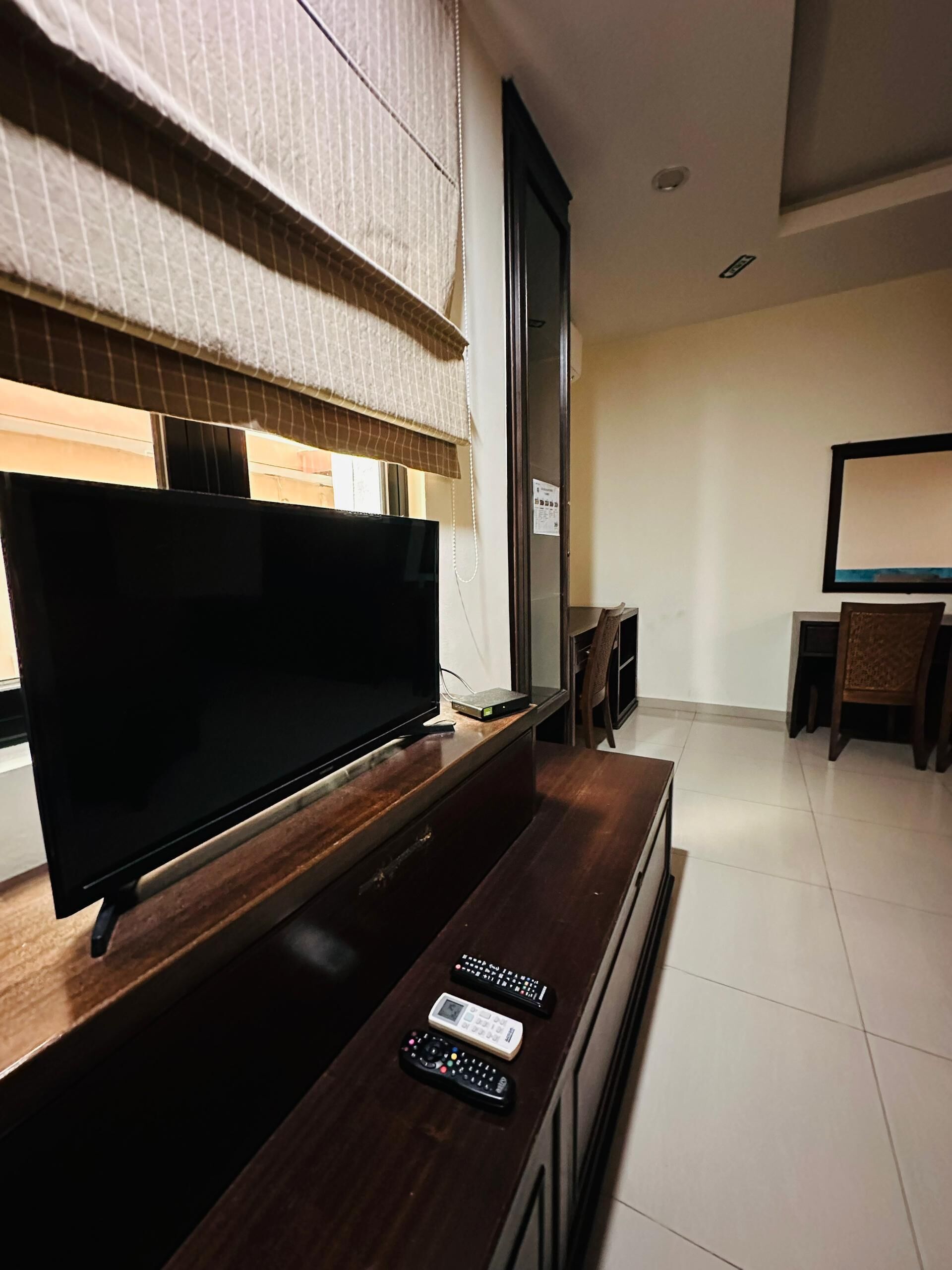 Una sala de estar moderna con una televisión de pantalla plana sobre un soporte de madera, una mesita con controles remotos y un comedor al fondo.