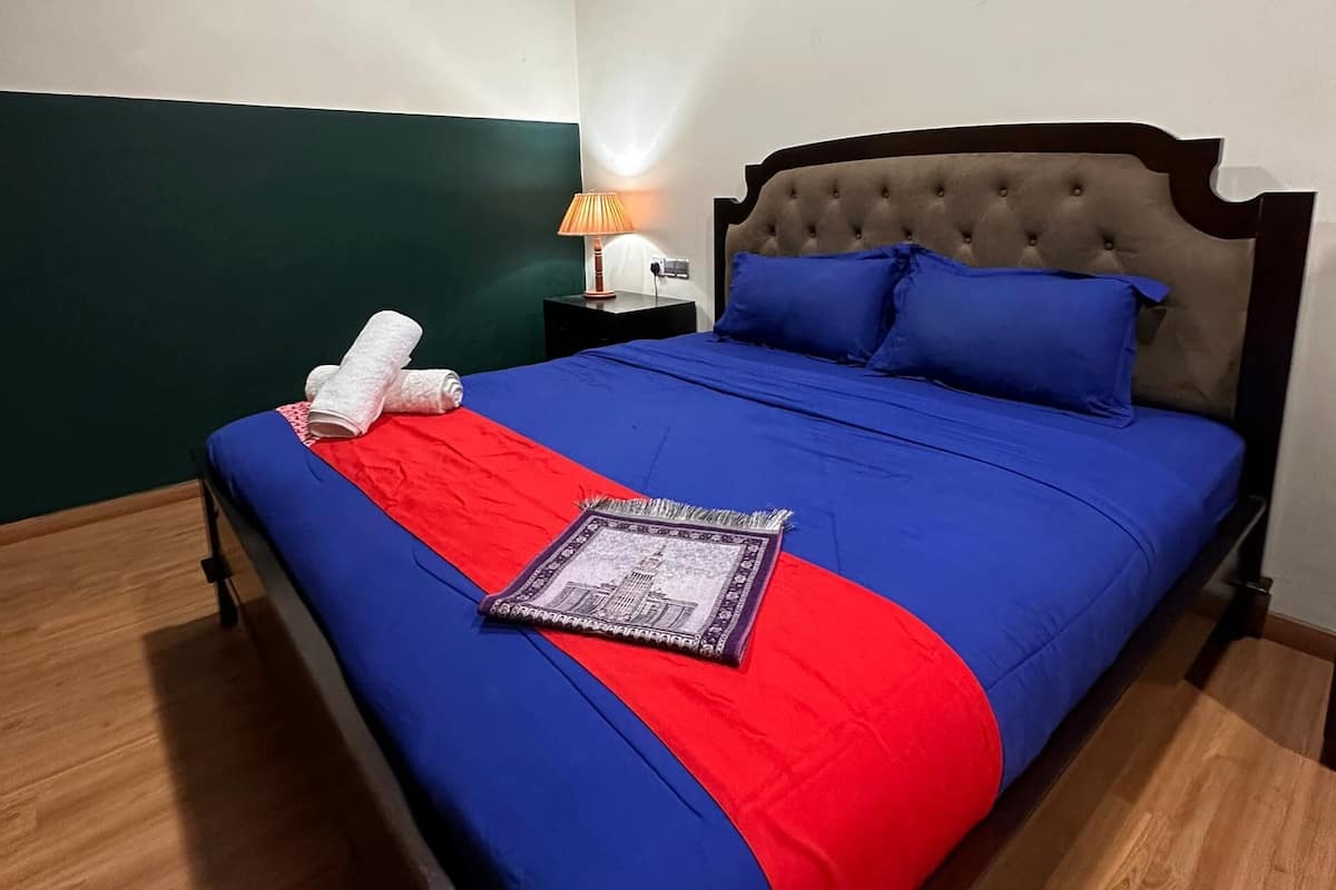 Un lit bien fait, avec une couvre-lit bleue et rouge, un coussin décoratif et une serviette pliée posée sur le côté.