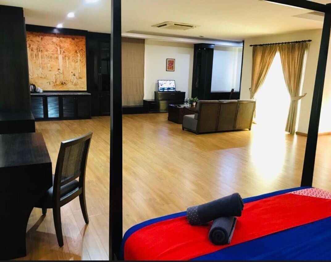 A hotel room with a wooden floor, a bed with a red and blue bedspread, a black chair, a television, and a wall with a large artwork.