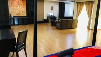A hotel room with a wooden floor, a bed with a red and blue bedspread, a black chair, a television, and a wall with a large artwork.