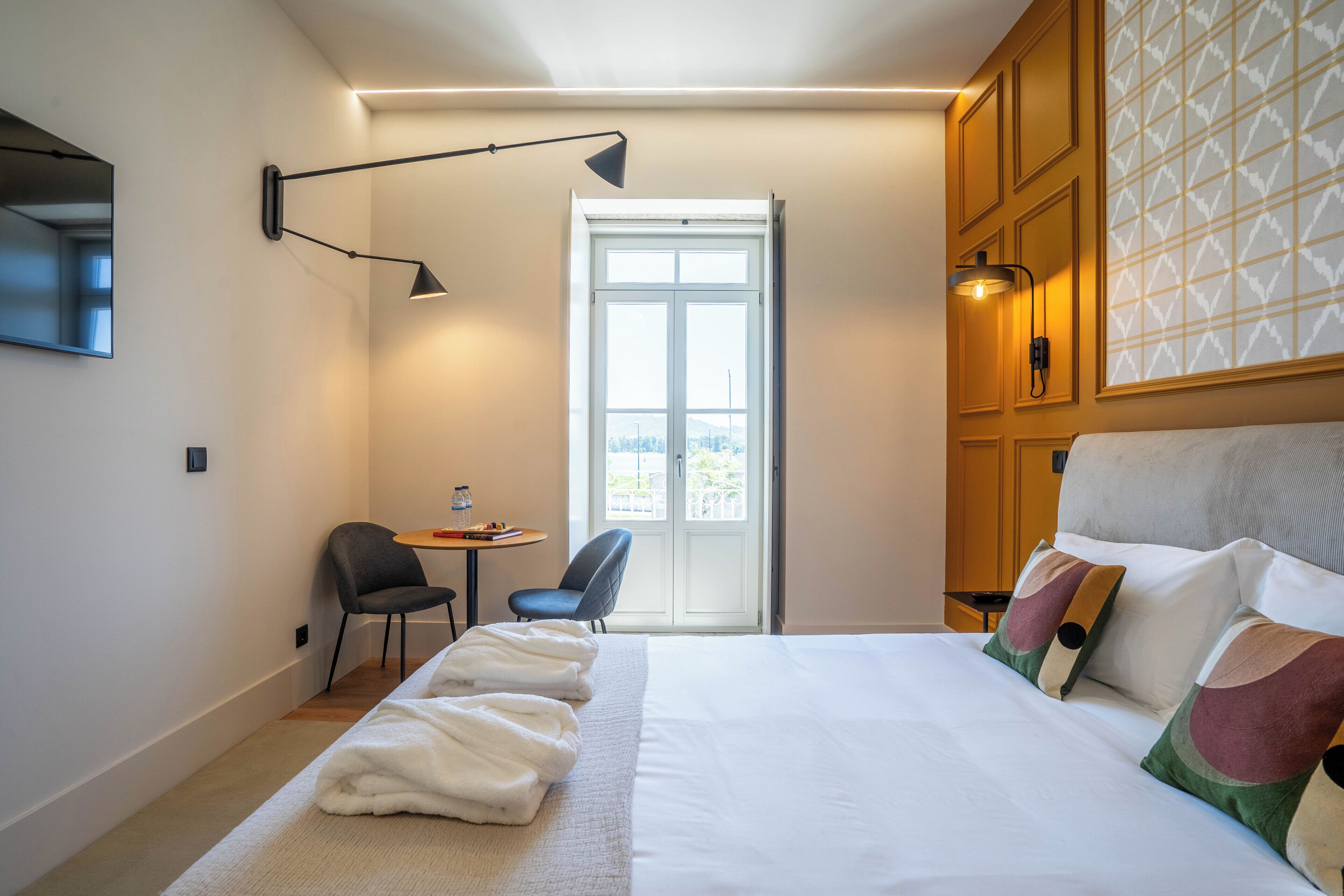 A hotel room with a large bed, a desk with two chairs, a window with a view, and a door.