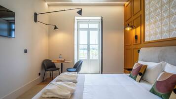 A hotel room with a large bed, a desk with two chairs, a window with a view, and a door.