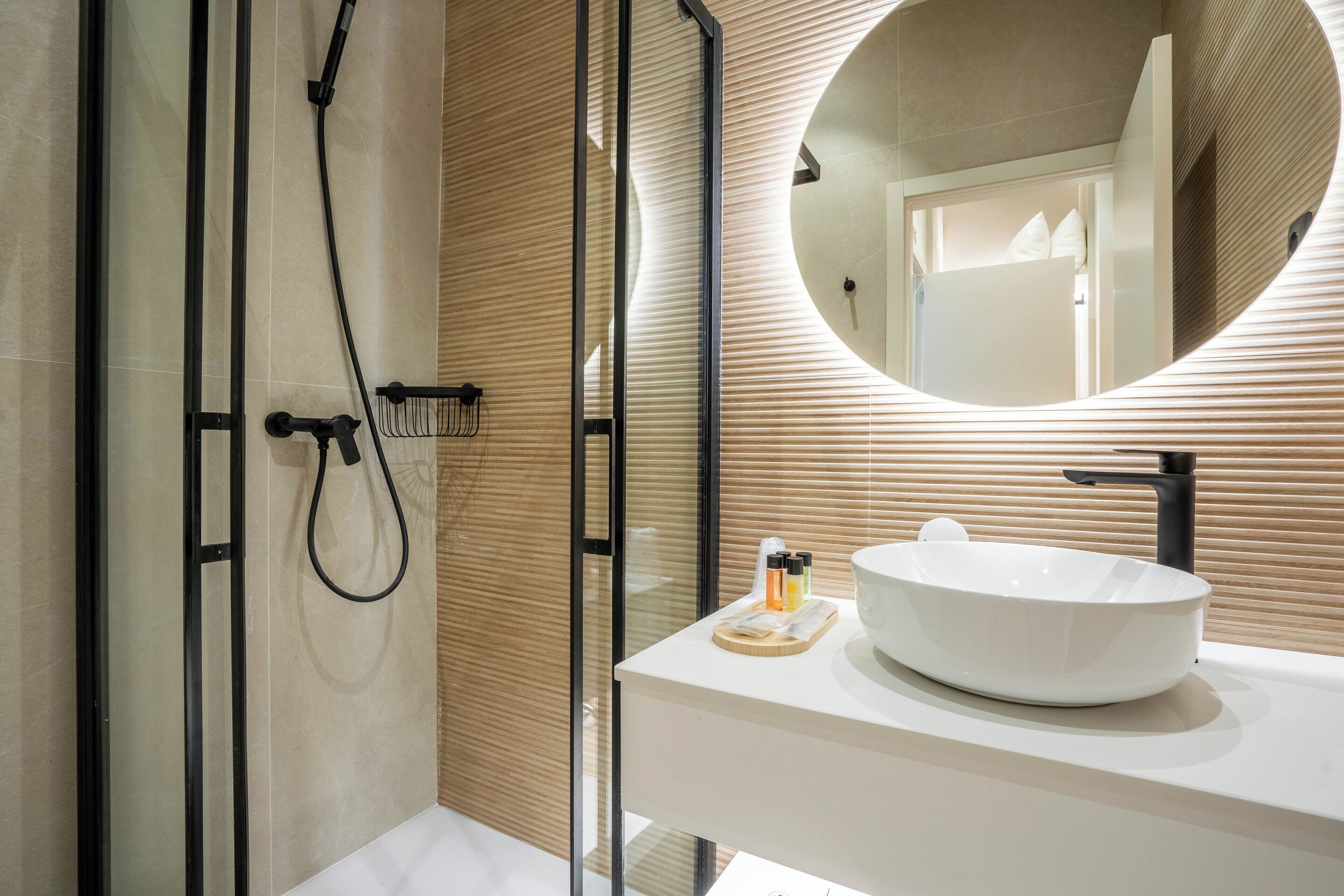 Un baño moderno con ducha acristalada, un espejo redondo y un lavabo blanco con lavamanos integrado.