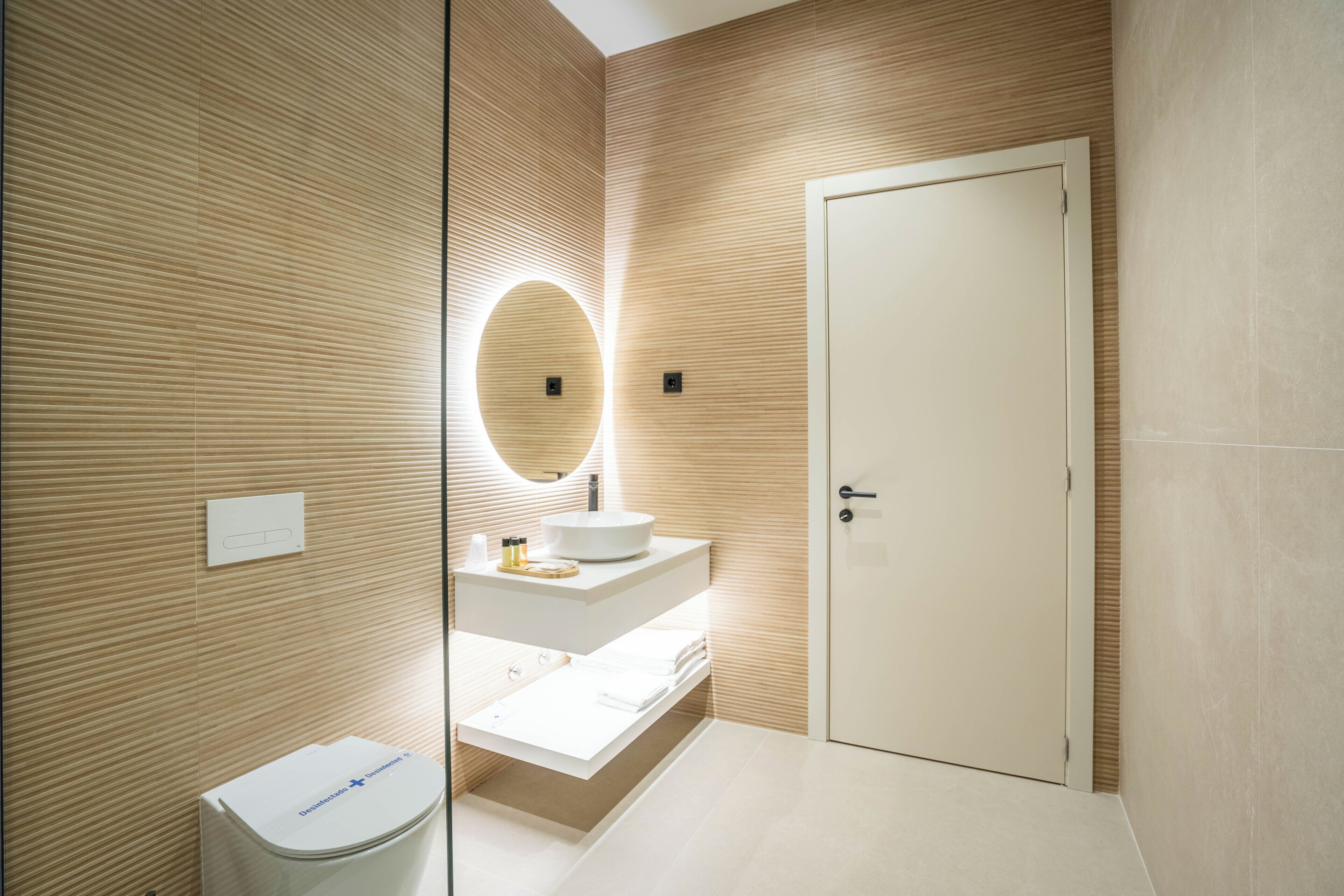 A modern bathroom with a wooden wall design, a round mirror, a white sink, and a toilet.