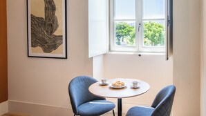 Une pièce avec une table ronde, deux chaises, une œuvre d’art encadrée et une fenêtre donnant sur des arbres.