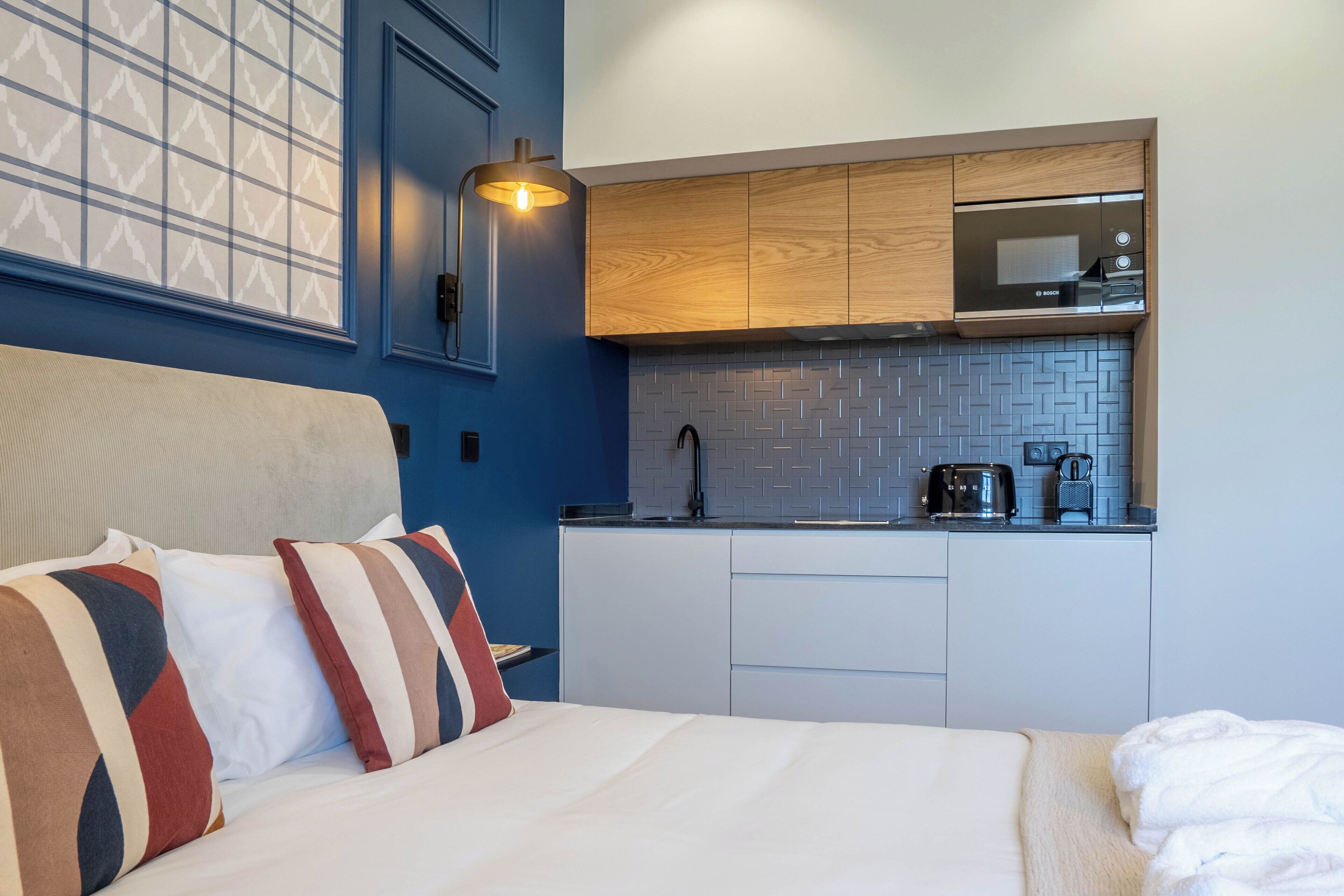 Ein kleines Hotelzimmer mit Bett, einer Küchenzeile inklusive Mikrowelle und einer Wandleuchte.