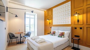 Une chambre d’hôtel moderne avec un grand lit, deux fauteuils, une petite table et une fenêtre ornée d’un motif géométrique.