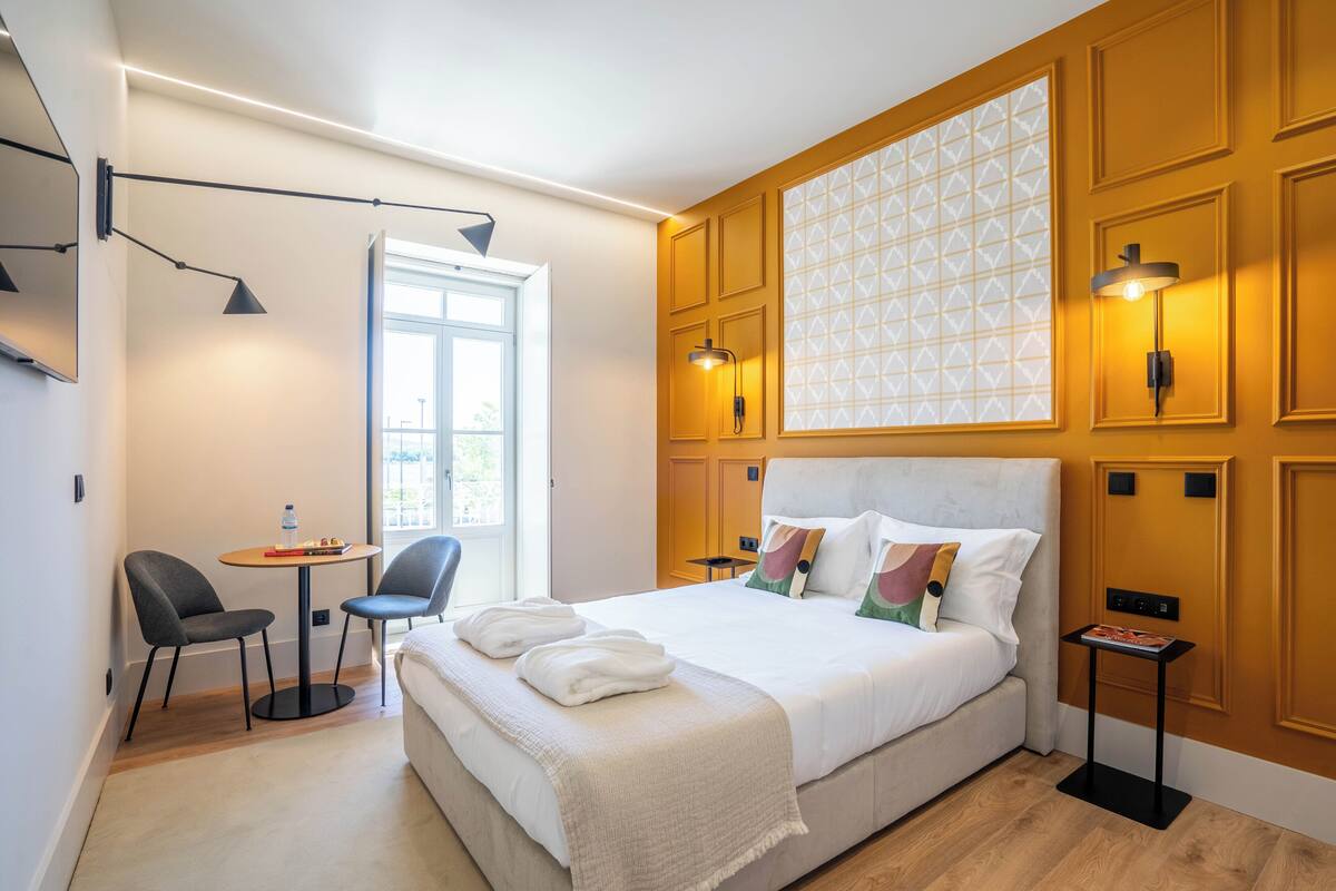 Ein modernes Hotelzimmer mit einem großen Bett, zwei Sesseln, einem kleinen Tisch und einem Fenster mit geometrischem Muster.