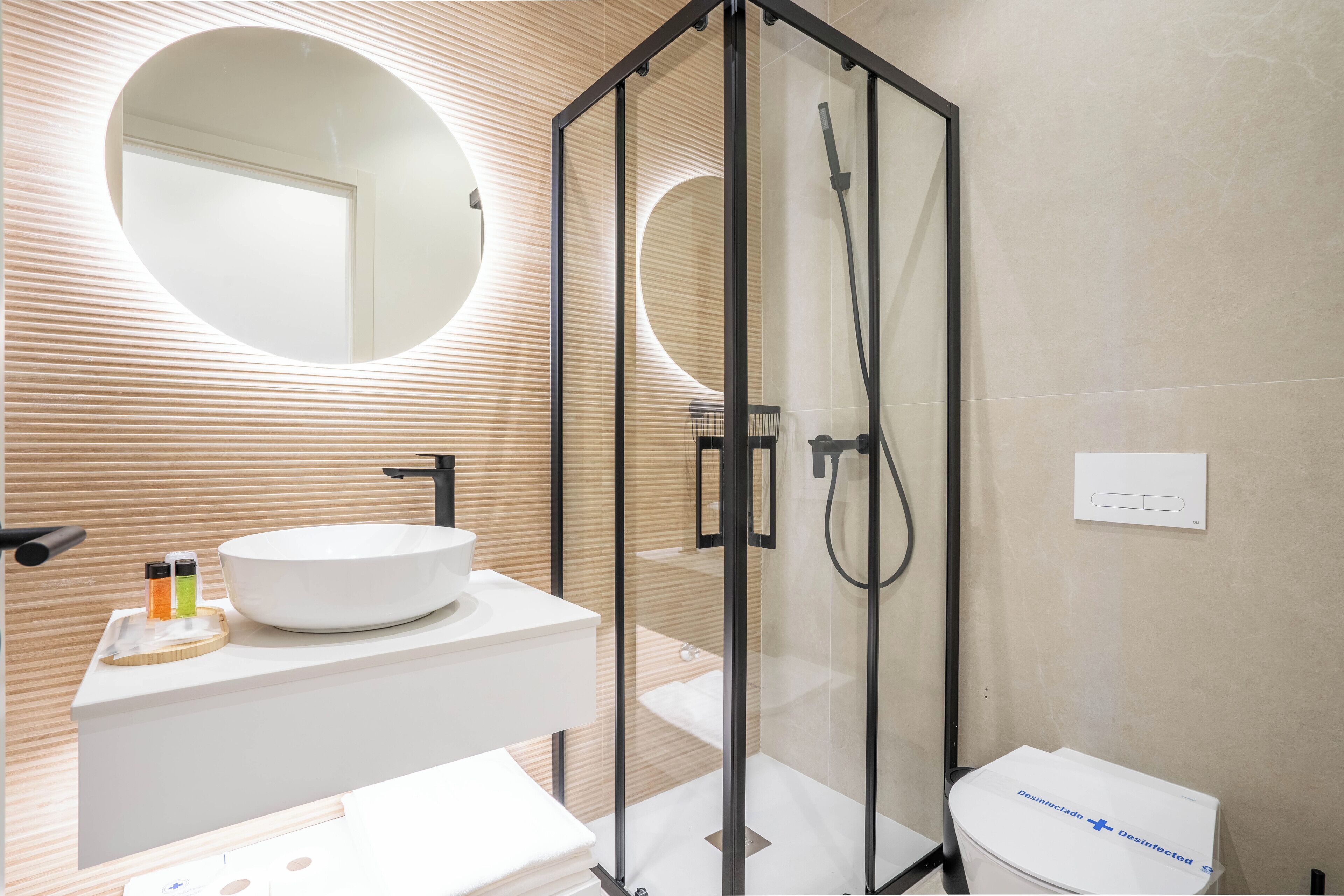 Een moderne badkamer met een douche in een glazen cabine, een ronde spiegel en een witte wastafel met een houten aanrecht.