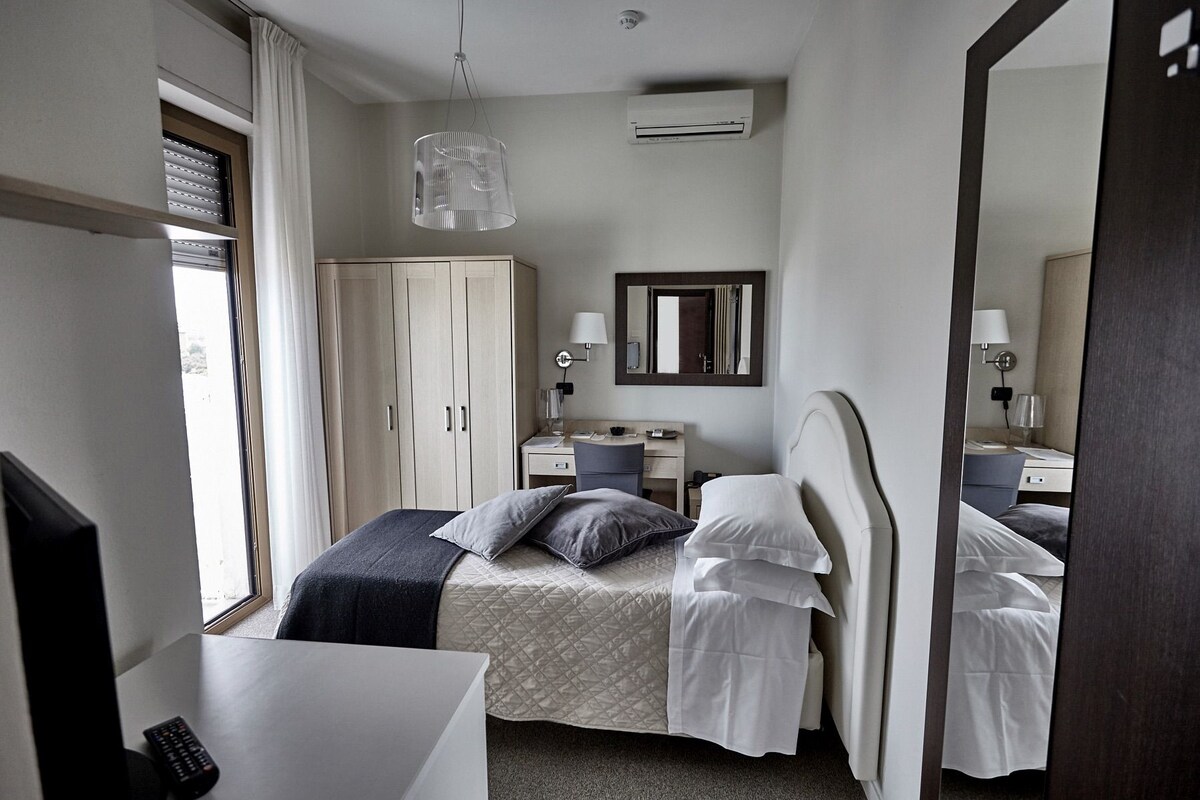 A hotel room with a bed, bedside tables, a desk, a wardrobe, a mirror, and an air conditioning unit.