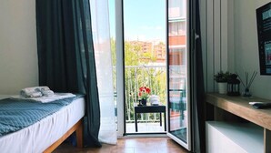 A bedroom with a bed, a desk, and a balcony with a view of buildings and trees.