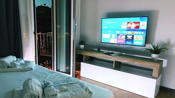 A modern hotel room with a bed, a TV on a wooden console, and a view of the city at night.