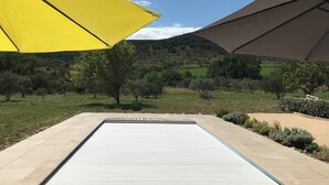 Una piscina amb un solàrium emblamat, envoltada per un jardí paisatjistic amb arbres i un paisatge amb turons al fons.