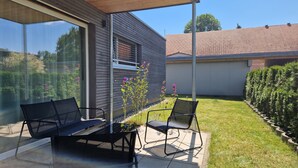 A patio with two chairs and a table, a wooden ceiling, a window, and a view of a house and garden.