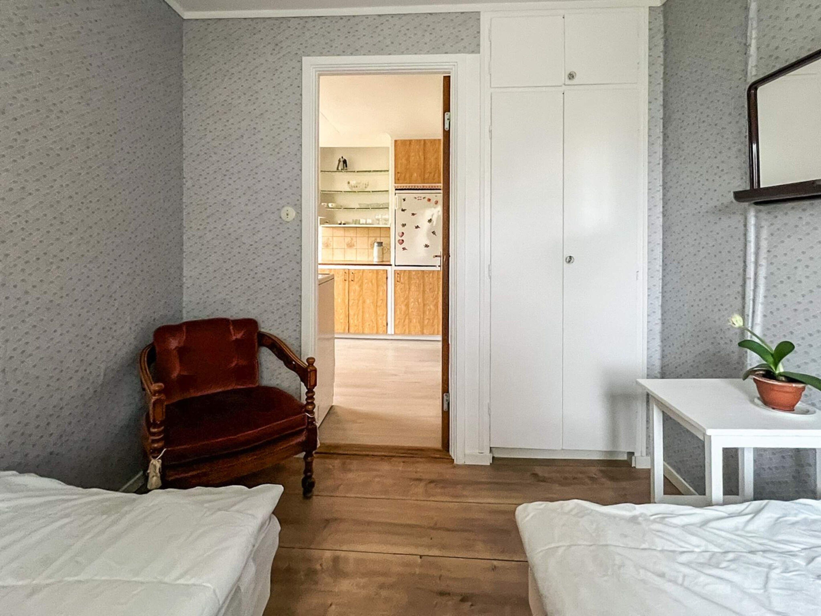 A room with two beds, a wooden chair, a small table with a plant, and a doorway leading to a kitchen.