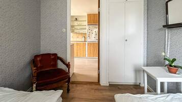 A room with two beds, a wooden chair, a small table with a plant, and a doorway leading to a kitchen.