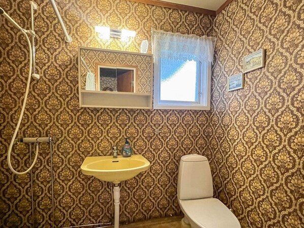Ein Badezimmer mit strukturiertem Wandpapier, einem Waschbecken, einer Toilette, einem Fenster mit Vorhang und einem Spiegel mit Ablage darüber.