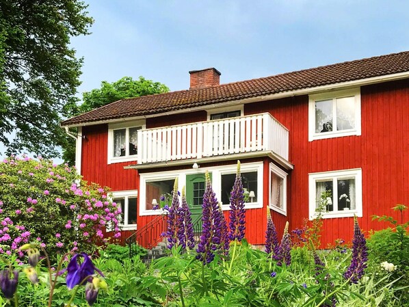 A red two-story house with a balcony, surrounded by lush greenery and purple flowers. - 6 Person Holiday Home in Lonashult (Lönashult)