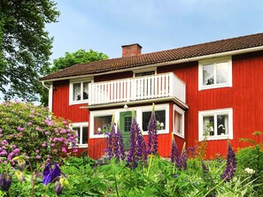 A red two-story house with a balcony, surrounded by lush greenery and purple flowers. - 6 Person Holiday Home in Lonashult (Lönashult)