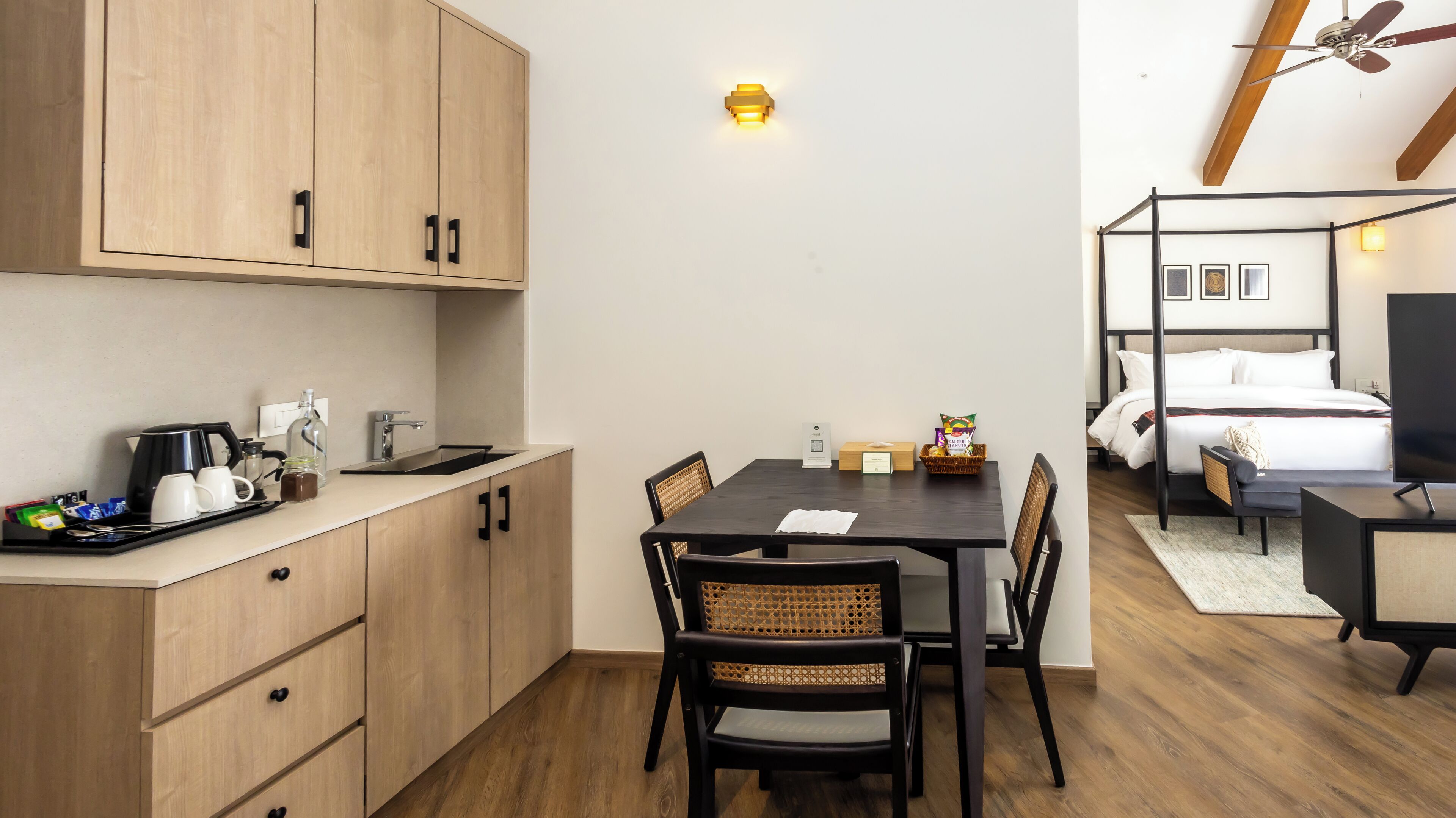 A compact hotel room with a kitchenette, a dining area with a table and chairs, a bed with a headboard, and a ceiling fan.