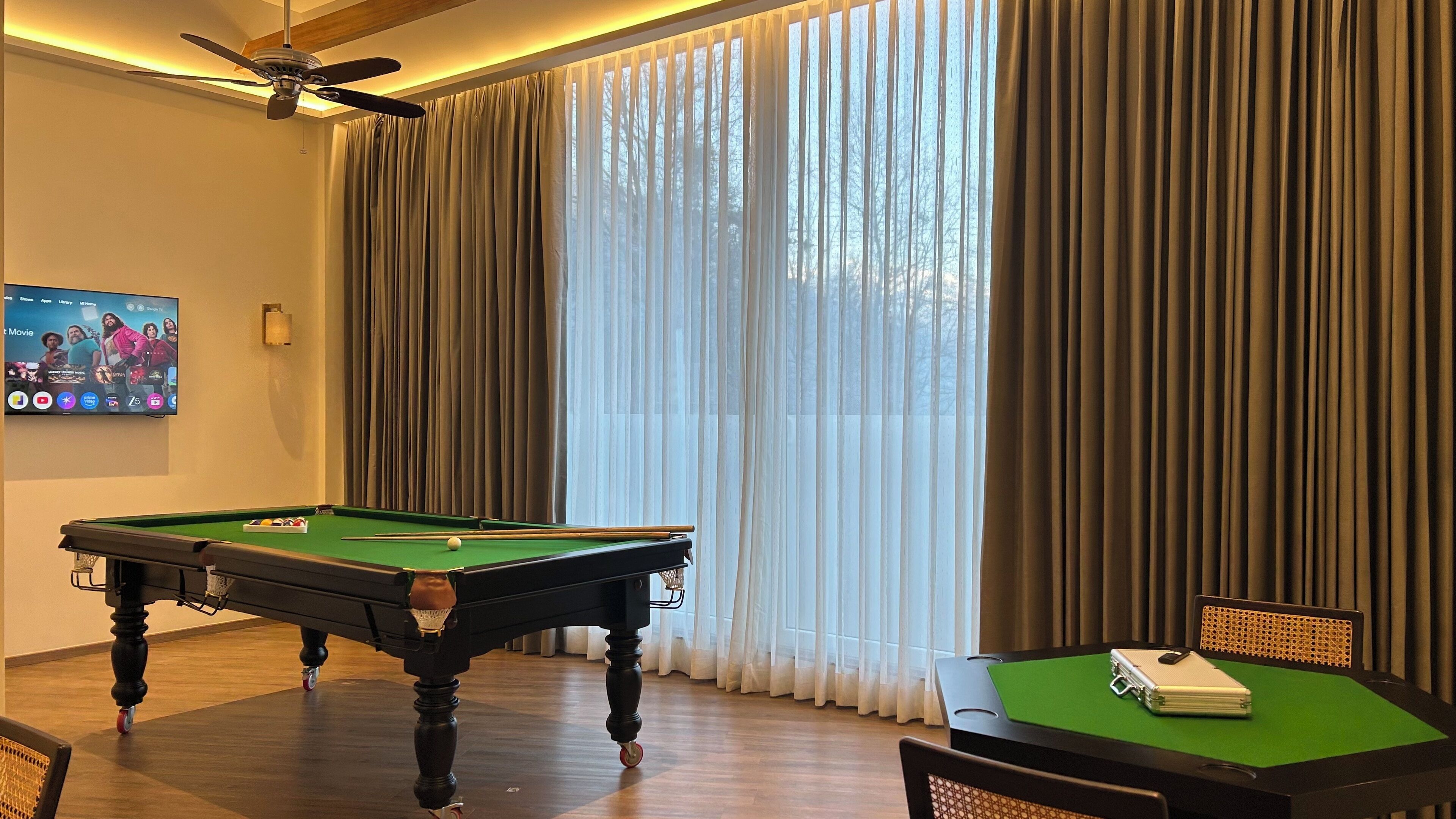 A pool table in a room with a ceiling fan, a television displaying a movie, and a window with curtains.