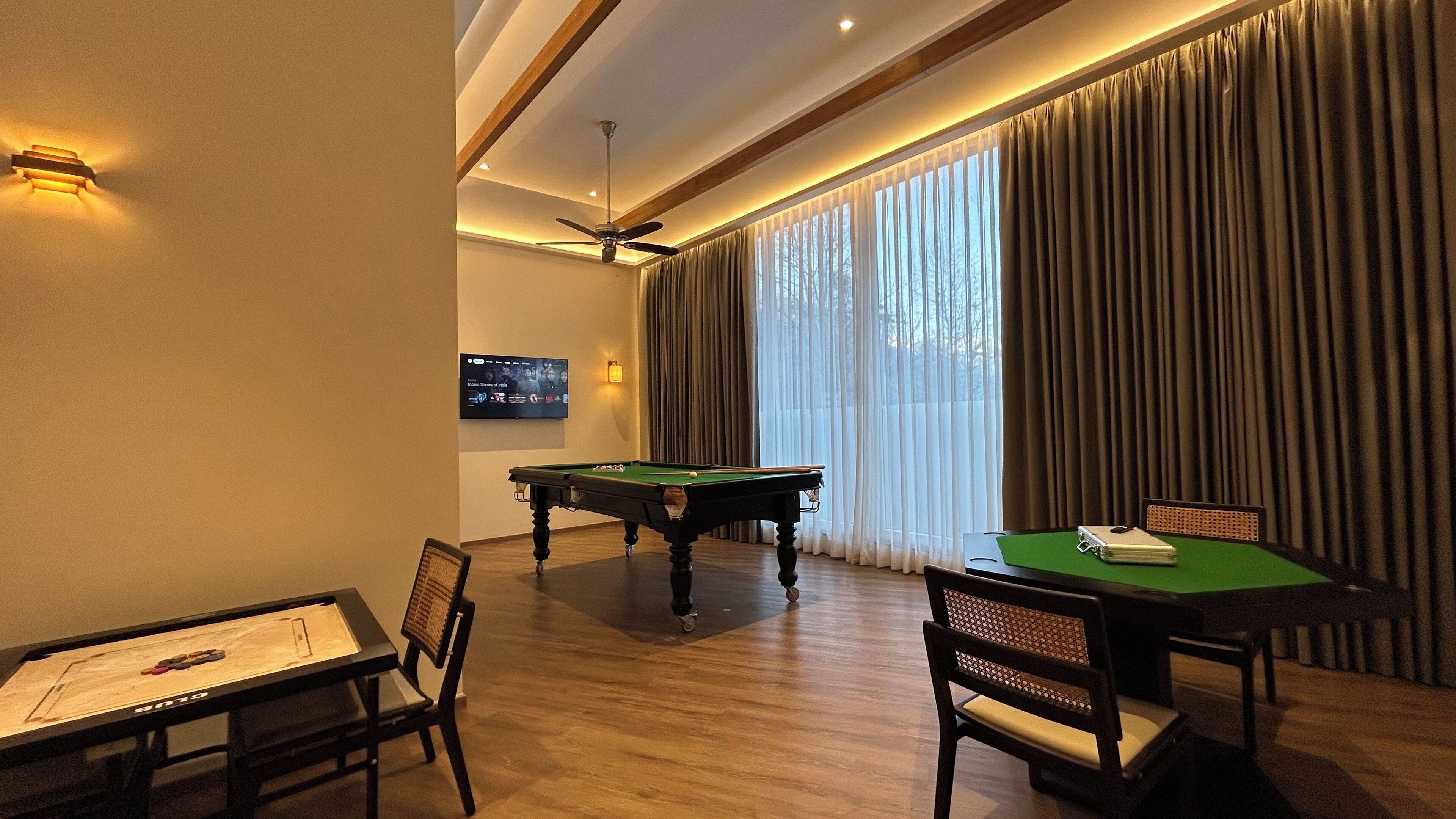 Une pièce équipée d’une table de billard, d’une table de snooker, d’un téléviseur fixé au mur et d’une grande fenêtre avec des rideaux.
