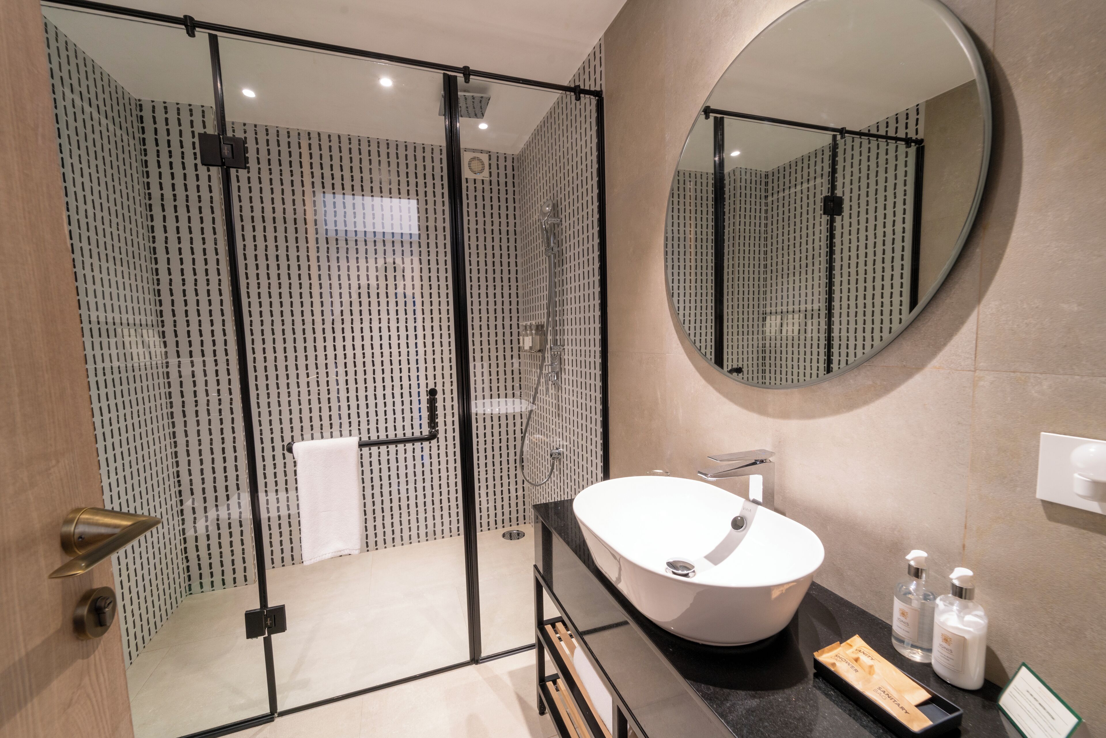 A modern bathroom with a glass-enclosed shower, a large round mirror, a white sink, and a black countertop.