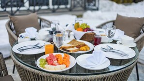 Una mesa de desayuno con platos de fruta, tostadas y bebidas, rodeada de sillas de mimbre y un paisaje nevado al exterior.