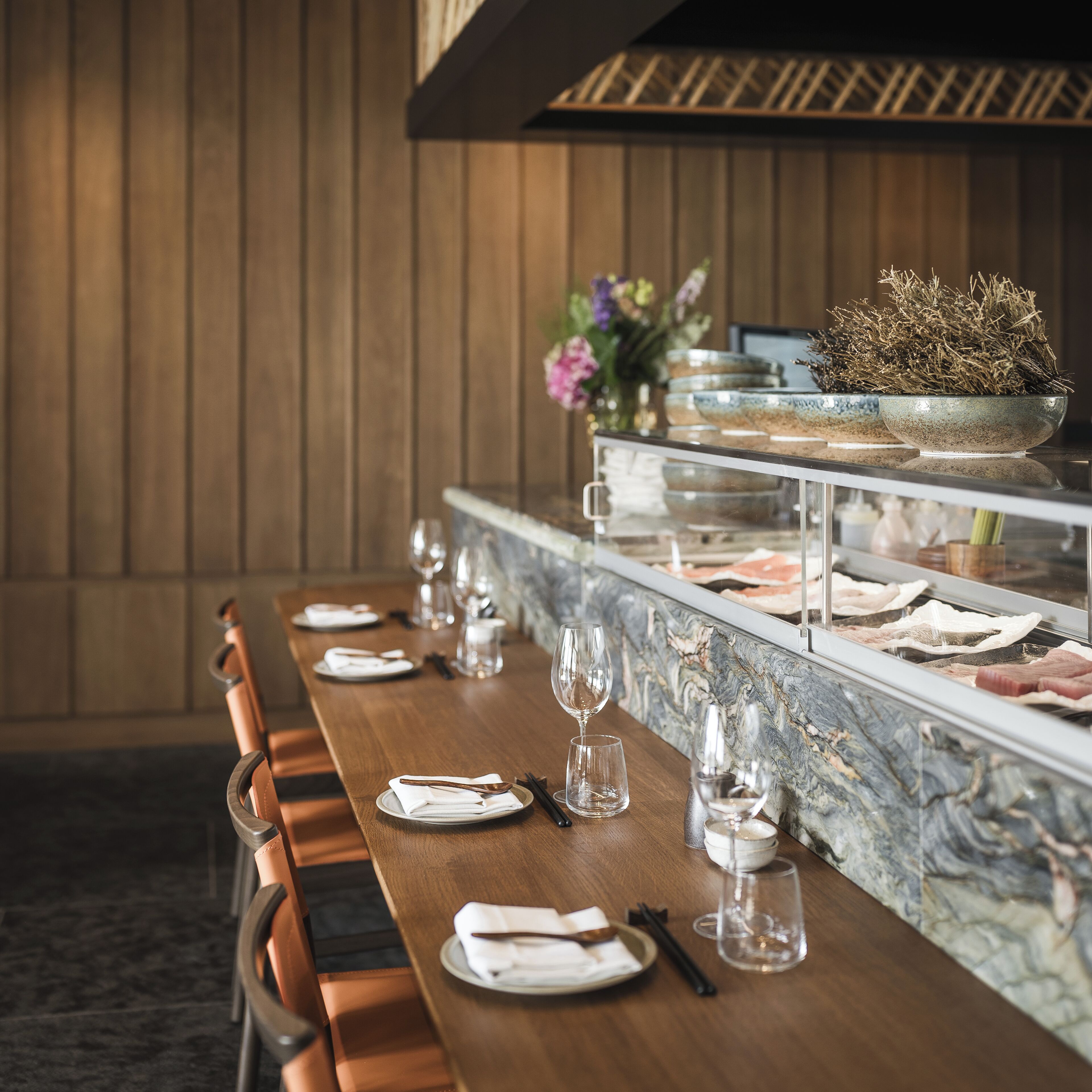 Una larga mesa de comedor de madera dispuesta con platos, cubiertos y vasos, con una encimera de mármol y un arreglo de plantas decorativo al fondo.