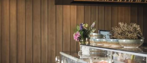 Una larga mesa de comedor de madera dispuesta con platos, cubiertos y vasos, con una encimera de mármol y un arreglo de plantas decorativo al fondo.