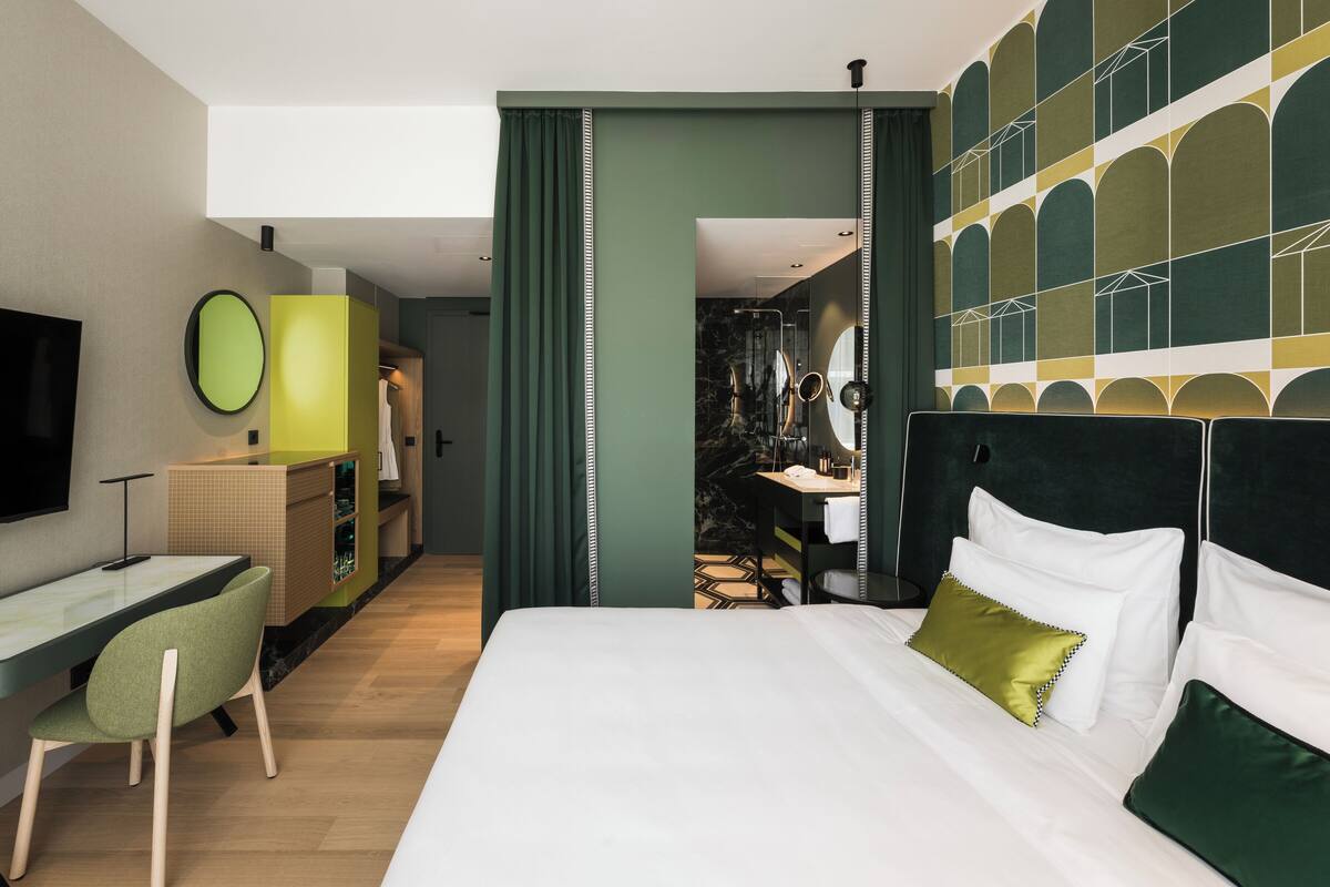 A hotel room with a bed, a desk, a chair, a TV, and a bathroom visible through an open door.