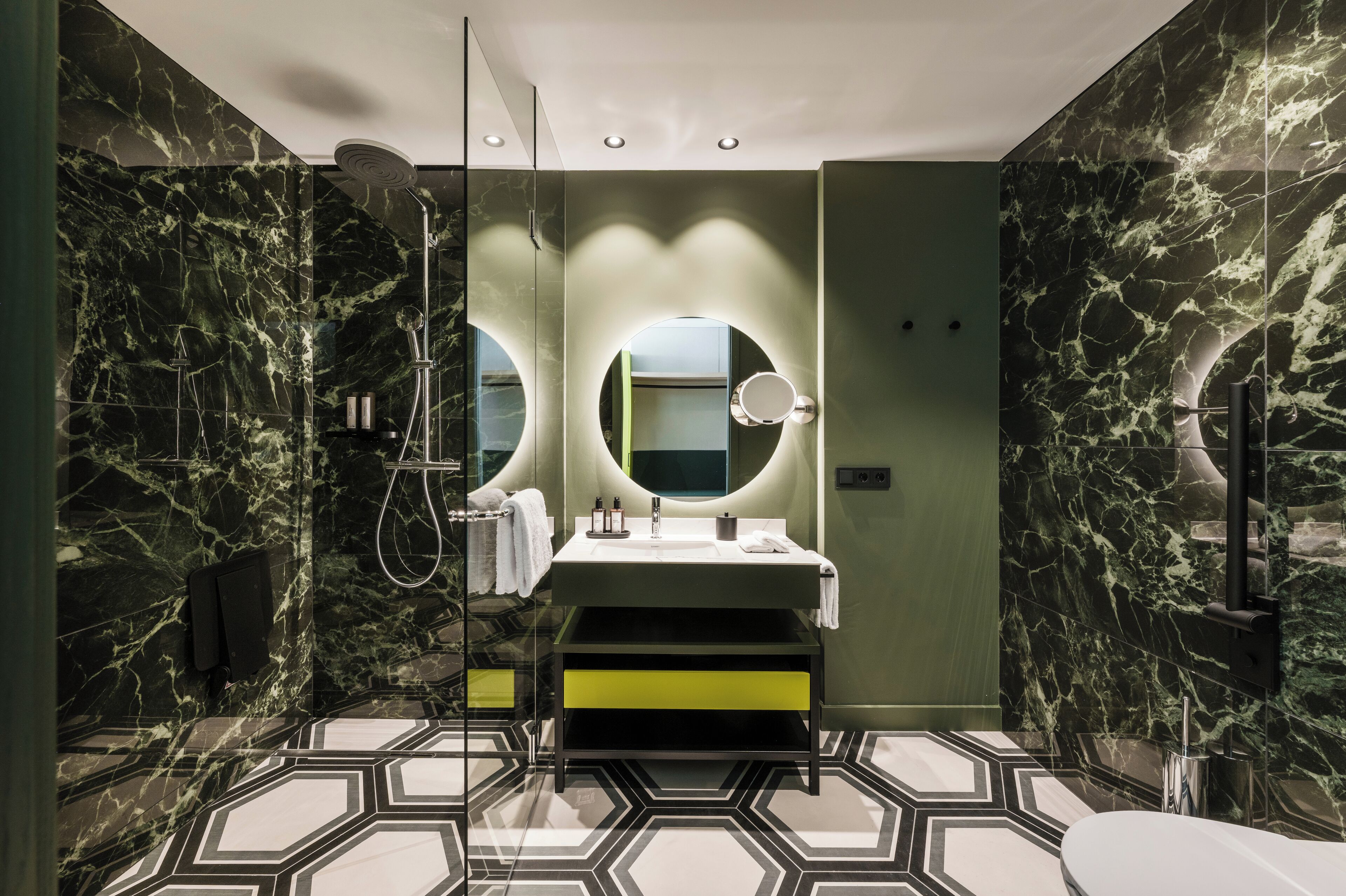 A modern bathroom with a marble shower, a round mirror, and a geometric patterned floor.
