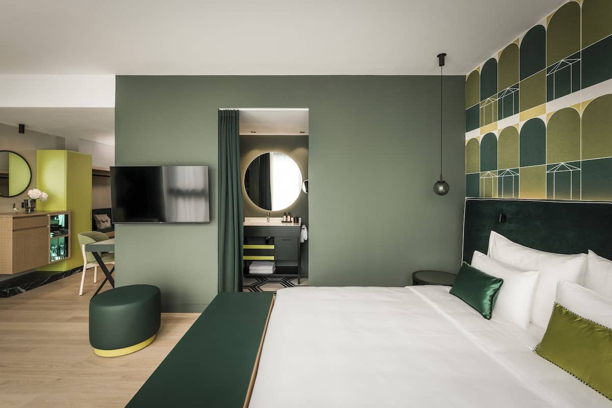 A modern hotel room with a large bed, a TV, a mirror, and a desk with a chair.