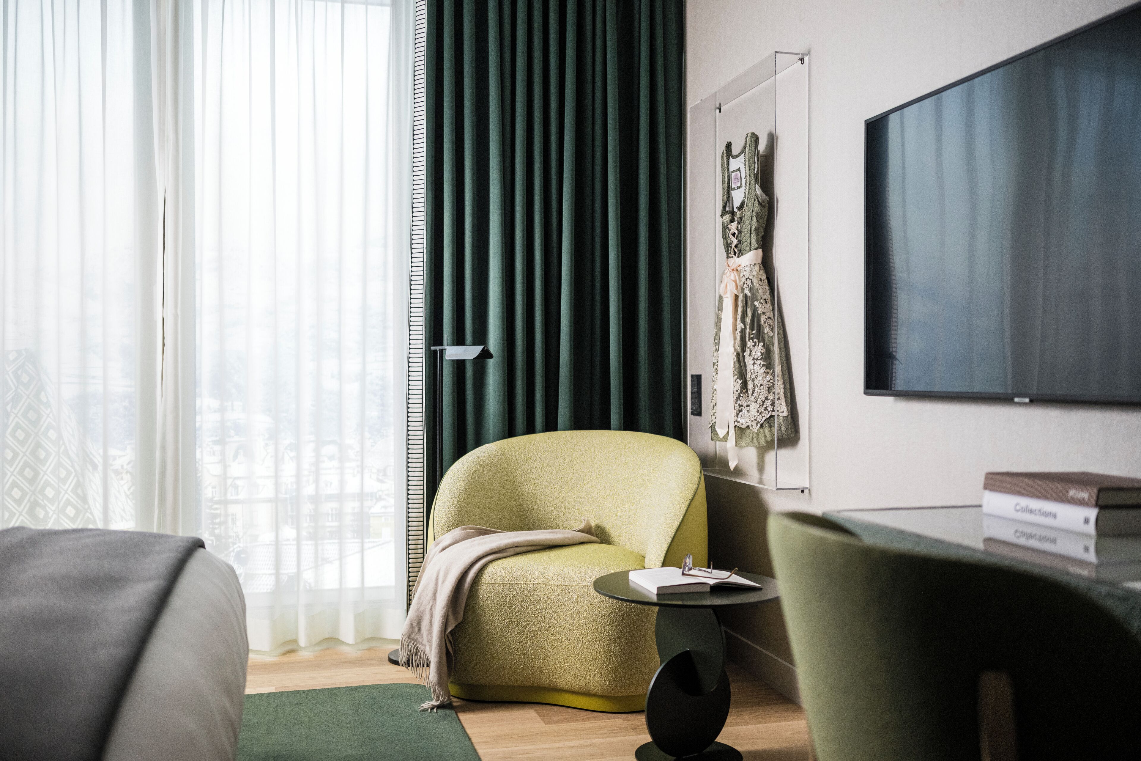 A modern hotel room with a green armchair, a small round table with a book, and a large flat-screen TV on the wall.