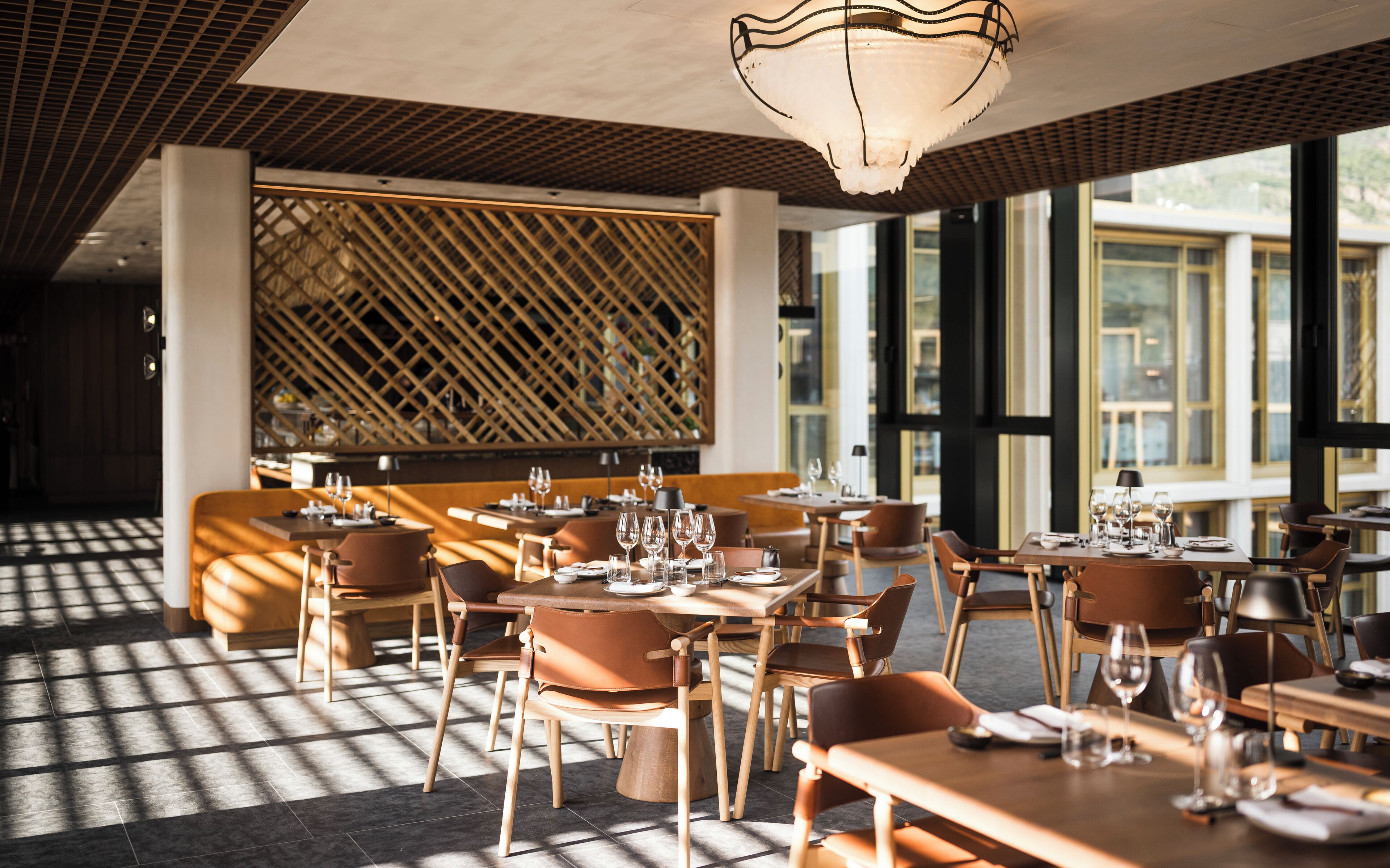 Un restaurante con mesas y sillas de madera, un muro con celosía y ventanas amplias.
