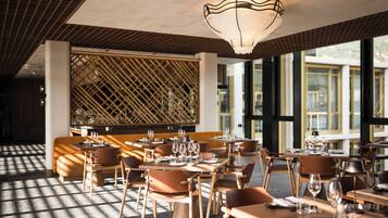 Un restaurante con mesas y sillas de madera, un muro con celosía y ventanas amplias.