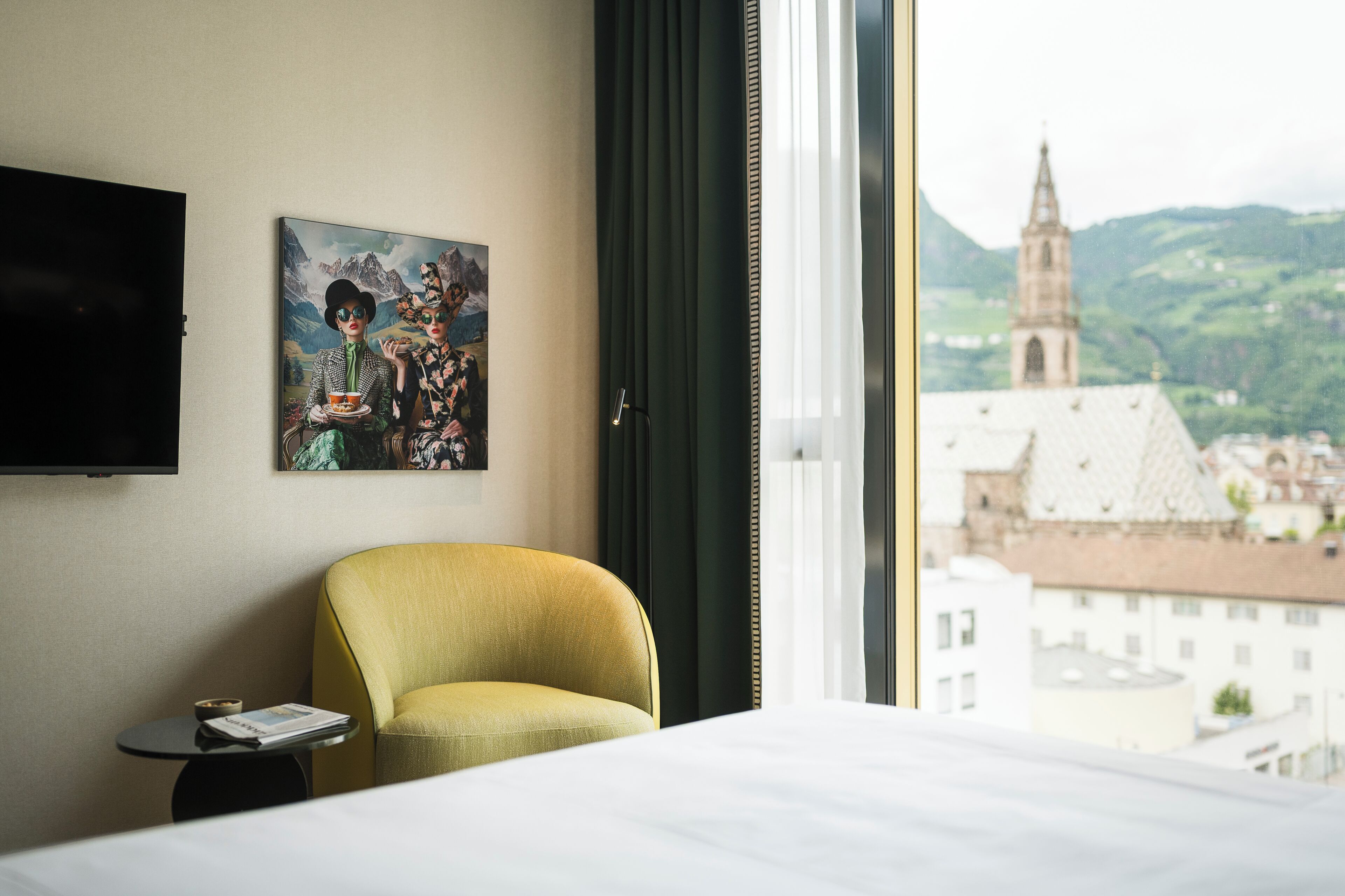 Habitación de hotel con cama, una silla, una mesita, un cuadro y vista a un paisaje urbano con una torre de iglesia.