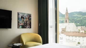 Habitación de hotel con cama, una silla, una mesita, un cuadro y vista a un paisaje urbano con una torre de iglesia.